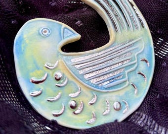 May include: A ceramic brooch shaped like a stylized bird with a light blue and pale yellow glaze. The bird features silver-toned metallic accents on the wing, tail feathers, and body, with a circular eye detail and a curved, rounded silhouette.