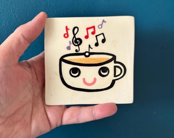 May include: Square ceramic coaster featuring a cartoon coffee cup with a smiling face. The cup is filled with a light brown liquid and surrounded by musical notes in red, black, and purple. The coaster is held by a hand.