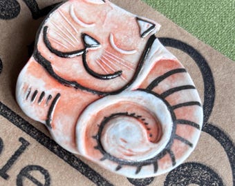 May include: A ceramic pin shaped like a curled-up cat with a peach and white glaze. The cat features black hand-painted outlines defining its closed eyes, whiskers, ears, and a striped tail that wraps around its body. The surface has a glossy finish.
