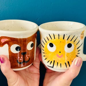 May include: Two ceramic mugs with quirky designs. One mug features a brown dog face, the other a yellow sun face. Both mugs have white speckled backgrounds and rounded handles. The mugs are held against a dark blue background.