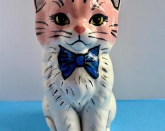 May include: A ceramic cat figurine with a pink head and a white body. The cat has yellow eyes, black whiskers, and a blue bow tie. The figurine is sitting upright.