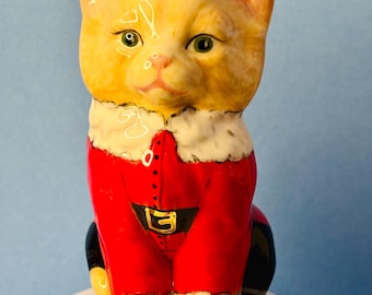 May include: A ceramic cat figurine wearing a red Santa Claus suit with a gold buckle. The cat has yellow fur, green eyes, and a white fur trim around the collar. The figurine is sitting on a white base.
