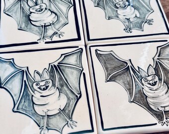 May include: A set of four square ceramic tiles, each featuring a hand-drawn illustration of a bat in flight. The bats are rendered in black and white with detailed wings, pointed ears, and visible fangs, set against a cream-colored background with a dark border.
