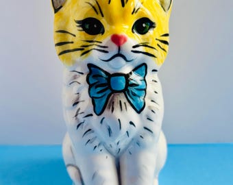 May include: A ceramic cat figurine with a yellow head and a white body. The cat has green eyes, a pink nose, and a blue bow tie. Black lines detail the cat's features. The figurine is sitting on a blue surface.