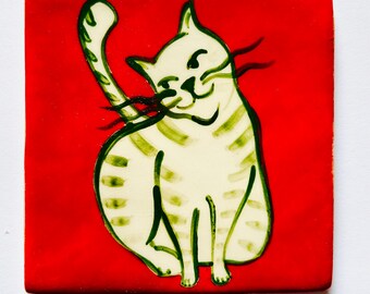 May include: Square ceramic tile with a red background featuring a cream-colored cat illustration. The cat is depicted with green stripes and a raised paw, and black whiskers. The tile is a decorative item.