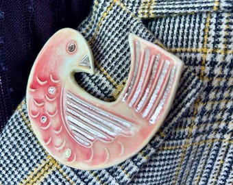 May include: A ceramic bird brooch in shades of pale pink and cream, featuring silver-toned accents on the wing and beak, with small embedded clear crystals along the body, pinned to a grey, white, and yellow plaid fabric with a dark purple texture.