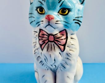 May include: A ceramic cat figurine with a light blue face and a white body. The cat has yellow eyes, a pink nose, and a pink bow tie. Black lines outline the cat's features. The figurine is sitting on a blue surface.