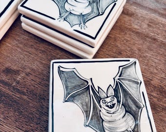 May include: A set of square ceramic tiles featuring a hand-drawn illustration of a bat with outstretched wings. The bat is rendered in shades of gray and black ink against a white background, enclosed within a thin black border on each tile.