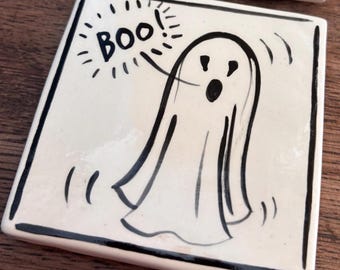 May include: A square ceramic coaster with a cream-colored surface and a black hand-drawn illustration of a ghost. The ghost has an open mouth and is surrounded by lines indicating movement. The text "BOO!" is written in black ink above the ghost.