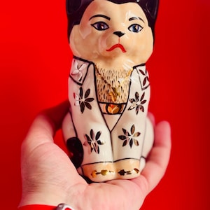 May include: A white ceramic cat figurine wearing a gold chain and a white and black patterned jacket. The cat has black ears and a black nose. The figurine is held in a hand with a red wristband.