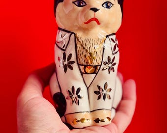 May include: A ceramic cat figurine featuring black ears, a cream-colored face, and a white body decorated with gold floral patterns. The cat wears a gold-colored chain necklace and has a painted face with blue eyes and red lips, presented in a seated pose.