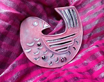 May include: A ceramic bird brooch in a soft pink hue with a matte finish. The bird features stylized wing and feather details highlighted with metallic silver accents. The design includes a curved tail and a small eye, showcasing a smooth, rounded shape.