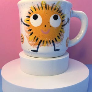 May include: A white ceramic mug featuring a cheerful sun design. The sun has a smiling face, arms, and legs, with orange and yellow rays. The mug is speckled and is displayed on a white pedestal, set against a pink backdrop.