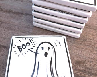 May include: A stack of square white ceramic coasters with black ink illustrations. Each coaster features a hand-drawn ghost with a black border and the text "Boo!" written in a speech bubble. The coasters have a glossy finish and smooth edges.