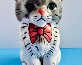 May include: A ceramic cat figurine with a gray and white coat, green eyes, and a red bow tie. The cat is sitting upright with its paws together. The figurine has a glossy finish and is set against a blue background.