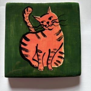 Può includere: Una piastrella quadrata in ceramica con un gatto tigrato arancione dipinto a mano con strisce nere su uno sfondo verde scuro uniforme. Il gatto è raffigurato in posizione seduta con la coda rivolta verso l'alto, con dettagli a linee nere sul viso.