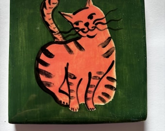 May include: A square ceramic tile featuring a hand-painted orange tabby cat with black stripes on a solid dark green background. The cat is depicted in a seated position with its tail curved upward, showcasing black line art details for its face and whiskers.