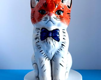 May include: A ceramic cat figurine with a red and white color scheme. The cat has a red head with black stripes and blue eyes. It wears a blue bow tie and sits on a white pedestal. The figurine is a decorative item.