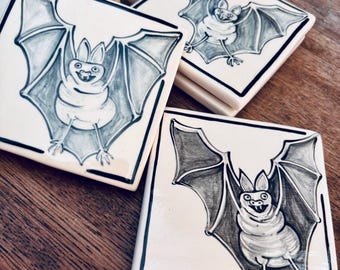 May include: A set of square ceramic coasters featuring a hand-drawn illustration of a bat with outstretched wings. The bat is rendered in shades of gray and black ink on a white background, with a thick black border framing the square tile design.