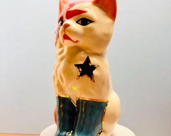 May include: A ceramic cat figurine with a cream-colored body, featuring a red lightning bolt design across one eye, a large black star on its chest, small gold star accents, and dark blue painted boots with gold trim on its front paws.
