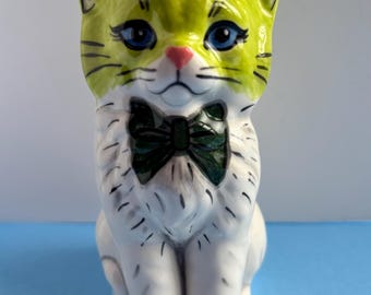 May include: A ceramic cat figurine with a lime green head and a white body. The cat has blue eyes, a pink nose, and a dark green bow tie. Black details outline the ears, whiskers, and facial features. The cat is sitting upright.