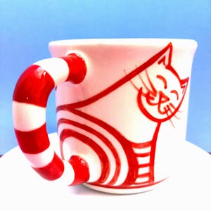 May include: A white ceramic mug with a red and white striped handle. The mug has a red cat design on the side, with a smiling face and striped body. The mug is on a white surface against a blue backdrop.