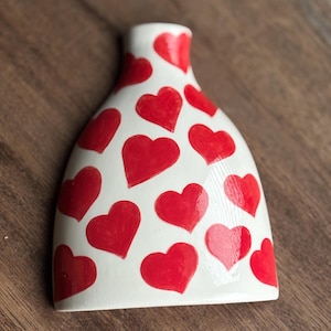 Puede incluir: Jarrón de cerámica blanca con un patrón de corazones. El jarrón está decorado con corazones rojos de varios tamaños.