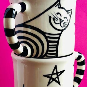 May include: Two white ceramic mugs with black and white designs. The top mug features a black and white cat with a striped tail. The bottom mug has a black and white star design.