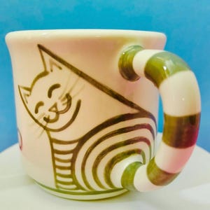 May include: A ceramic mug featuring a cat design. The off-white mug has a smiling cat illustration in brown. The handle is striped with green, white, and pink. The mug is a standard size for tea or coffee.