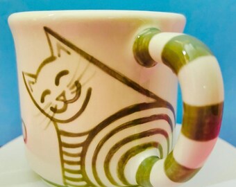 May include: A cream-colored ceramic mug featuring a hand-painted brown line drawing of a smiling cat with a striped body. The mug has a thick, curved handle decorated with alternating horizontal bands of olive green and cream.