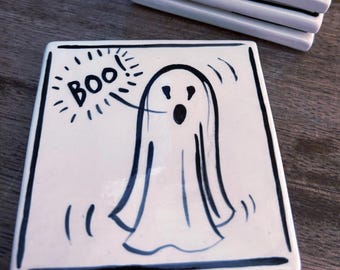 May include: A square white ceramic coaster featuring a hand-drawn black ink illustration of a ghost. The ghost has a surprised expression with an open mouth and is accompanied by a speech bubble containing the word Boo in black lettering.