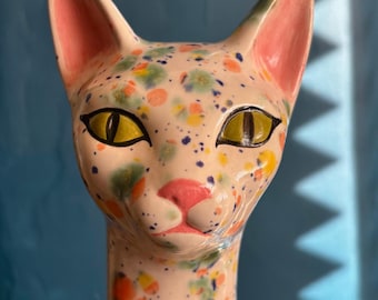 May include: A ceramic cat figurine with a speckled design. The cat's head is painted with a light pink base and colorful spots of orange, green, yellow, and blue. It has yellow eyes, black eyeliner, and a pink nose and inner ears. The figurine has a long neck.