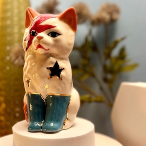 Può includere: Figurina di gatto in ceramica bianca con un design a stella rosso, nero e oro. Il gatto indossa stivali blu con accenti dorati.