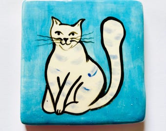 May include: A square ceramic tile featuring a hand-painted illustration of a white cat with faint grey stripes. The cat is depicted in a seated position against a vibrant, textured sky-blue background. The design uses bold black outlines.