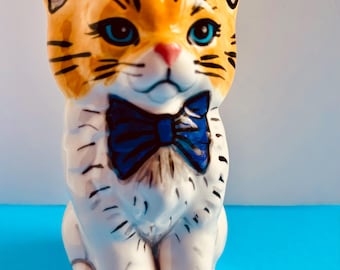May include: A ceramic cat figurine with orange and white fur, blue eyes, and a blue bow tie. The cat is sitting upright with its paws together. Black stripes accent the orange fur. The figurine is on a blue surface.