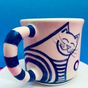 May include: A ceramic mug featuring a playful cat design. The mug is white with a blue cat illustration and a blue and white striped handle. The cat has a smiling face and is wearing a striped shirt. The mug is on a white surface with a blue backdrop.