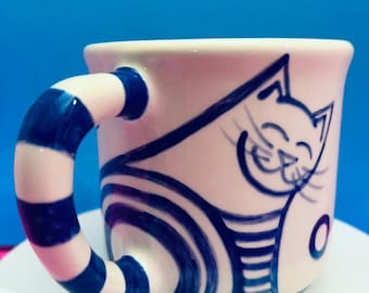 May include: A cream-colored ceramic mug featuring a hand-painted dark blue cat illustration. The cat has a smiling face and striped body. The mug handle is decorated with alternating cream and dark blue horizontal stripes, matching the cat design.