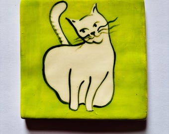 May include: Square ceramic tile with a hand-drawn illustration of a white cat on a bright green background. The cat is depicted with black outlines and a curved tail, giving it a playful appearance. The tile has a glossy finish.