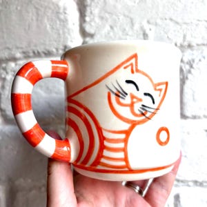 Può includere: Una tazza in ceramica color crema con un disegno a linee arancioni dipinto a mano che raffigura un gatto sorridente con striature. La tazza presenta un manico curvo decorato con strisce orizzontali arancioni e bianche e un piccolo cerchio arancione.