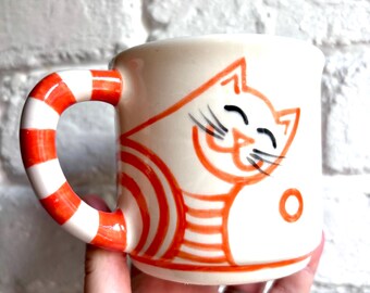 May include: A cream-colored ceramic mug featuring a hand-painted orange line drawing of a smiling cat with striped markings. The mug has a curved handle decorated with horizontal orange and white stripes and a small orange circle on the side.