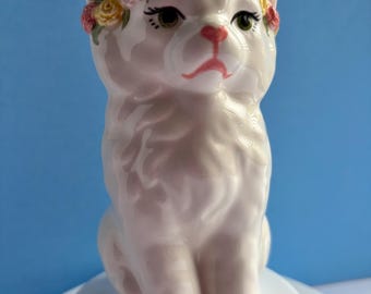 May include: A white ceramic cat figurine featuring a glossy finish, painted green eyes, and a pink nose. The cat is adorned with a sculpted floral crown consisting of small pink and yellow roses resting between its ears on its head.