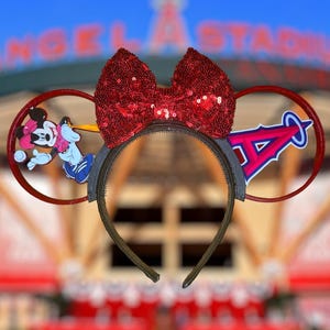 May include: A red sequined bow headband with a gray denim-like band. The ears feature a Minnie Mouse and Donald Duck graphic on one side and an Angels baseball team logo on the other. The background is a blurred image of a stadium.