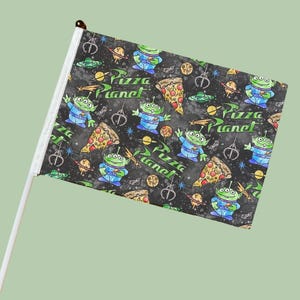 May include: A hand-held flag featuring a repeating pattern of green aliens, pizza slices, and the words "Pizza Planet" on a black background. The flag is attached to a white pole with a brown tip.