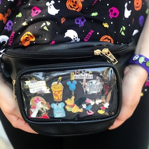 May include: Black fanny pack with a clear front pocket displaying Disney pins. The fanny pack has a gold zipper and is held by a person wearing a black shirt with a Halloween-themed pattern of pumpkins, ghosts, and skulls.