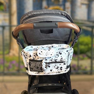 May include: A stroller organizer with a black and white Mickey Mouse pattern. The organizer has multiple pockets, including a mesh pocket and a zippered pocket. The stroller handle is brown leather. The background is blurred, showing a castle.