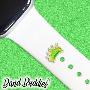 May include: A green and gold crown-shaped charm is attached to a white watch strap. The words "Band Buddies" are written in a cursive font at the bottom of the image. The background is a glittery green.