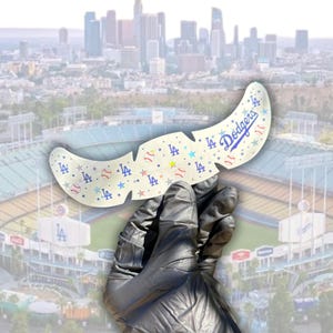 May include: A cream-coloured, feather-shaped accessory featuring the Los Angeles Dodgers logo and baseball-themed graphics. The design includes blue stars, baseballs, and the letters "LA". The Dodgers text is in blue script.