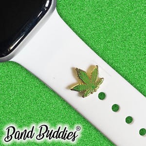 May include: A gold and green cannabis leaf charm attached to a white smartwatch band. The band is on a green textured background. The words "Band Buddies" are written in a cursive font at the bottom of the image.