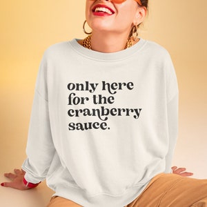 May include: A white sweatshirt with the text "only here for the cranberry sauce." printed in black. The model is wearing the sweatshirt and smiling.