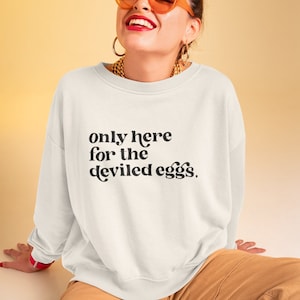 Può includere: Una felpa bianca con la scritta "only here for the deviled eggs." stampata in nero. La modella indossa la felpa e sorride.